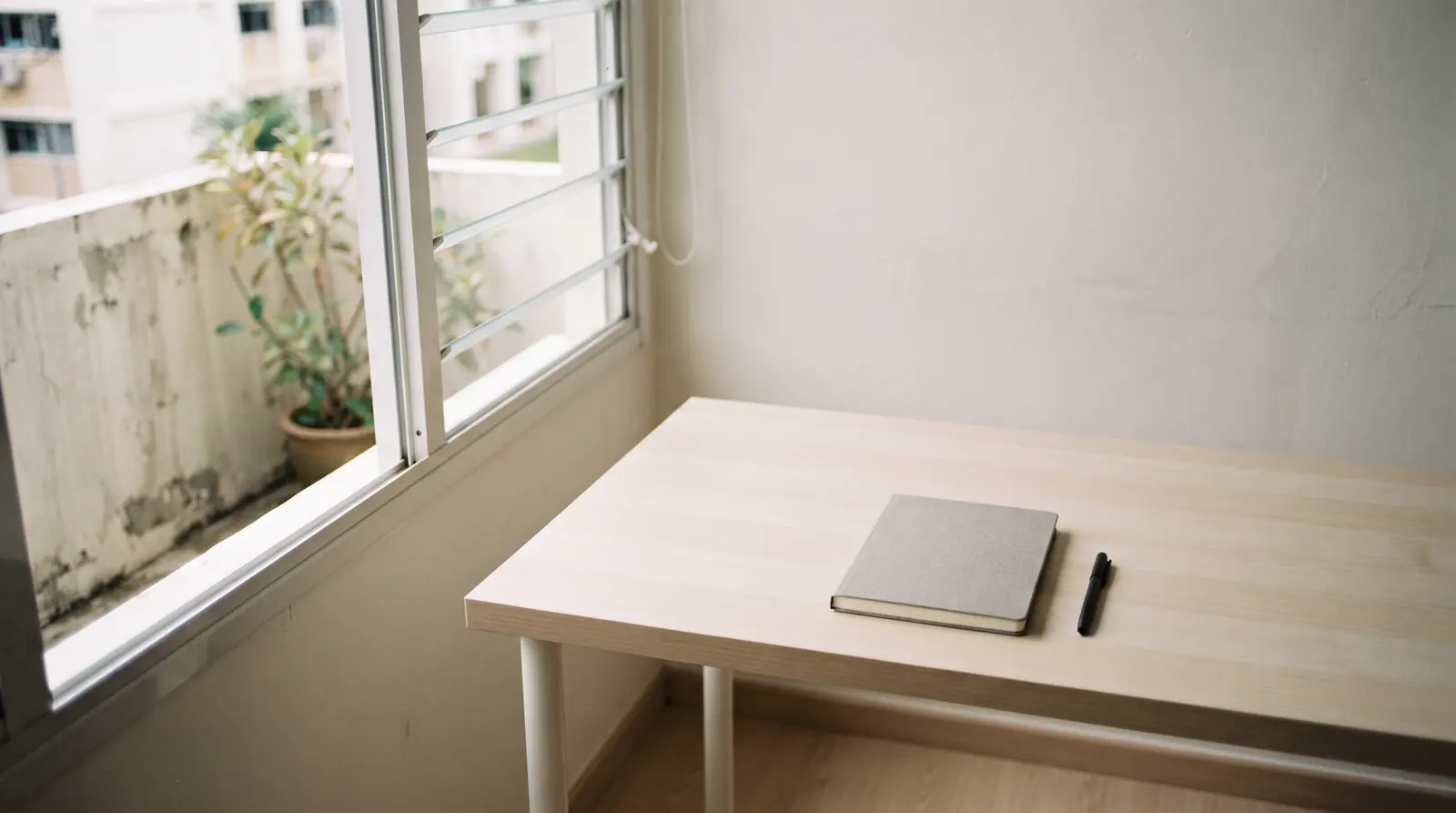 A pale desk with a closed unmarked notebook beside a window